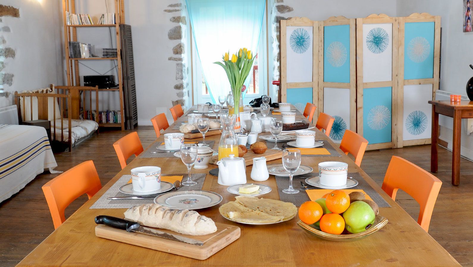 Een eetruimte met een houten tafel gedekt voor het ontbijt, oranje stoelen en verschillende ontbijtartikelen, zoals brood, fruit en drankjes.