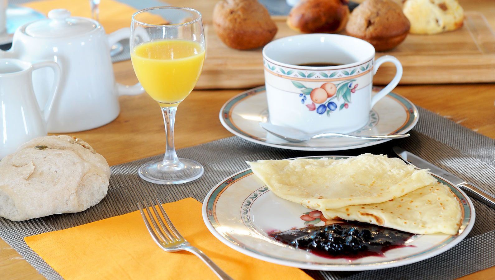 Aamiaispöytä, jonka päällä on lautanen täynnä kreppejä, lasi appelsiinimehua, kuppi kahvia ja leivonnaisia.