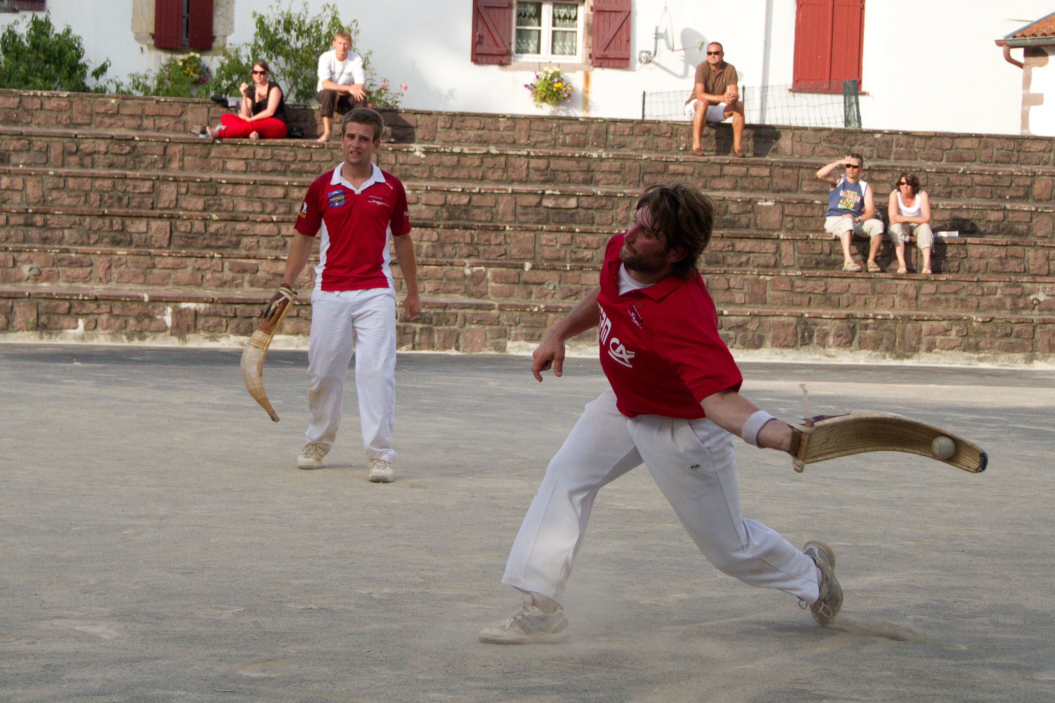 Partido de críquet en desarrollo, con jugadores uniformados de rojo y blanco en un campo de hormigón.