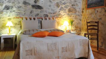 Un dormitorio con pared de piedra, una cama con ropa de cama blanca y almohadas naranjas, dos lámparas de noche y una silla de madera.