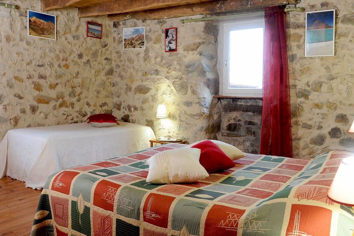 Une chambre rustique aux murs de pierre, avec deux lits recouverts de courtepointes, une fenêtre ornée de rideaux rouges et une lampe sur une table de chevet.