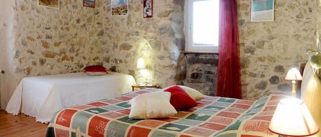 A rustic room with stone walls, two beds with quilts, a window with red curtains, and a lamp on a bedside table.