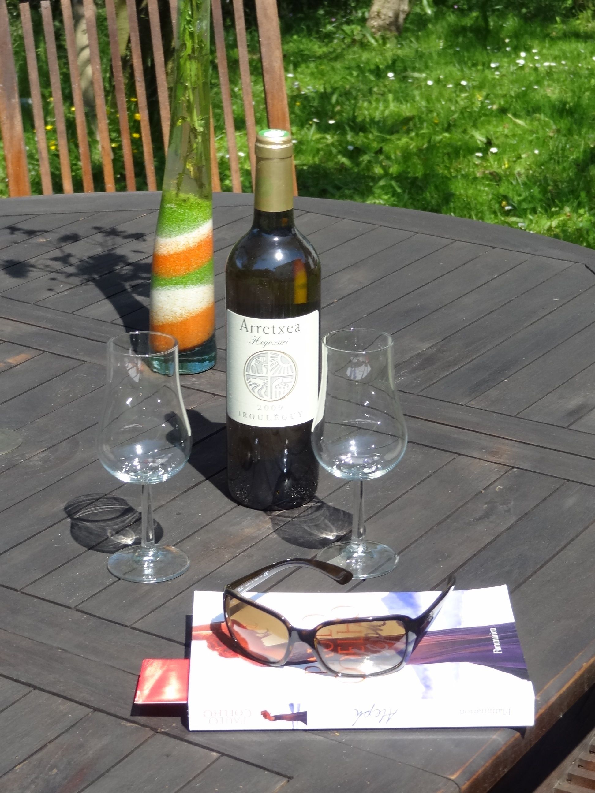 Una botella de vino Arretxea, dos copas y un par de gafas de sol sobre una mesa de madera al aire libre.