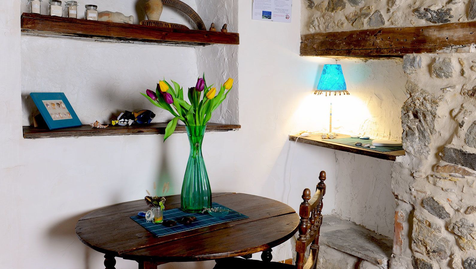 Een rustieke kamer met een ronde houten tafel, een groene vaas met bloemen, een blauwe lamp en stenen muren.