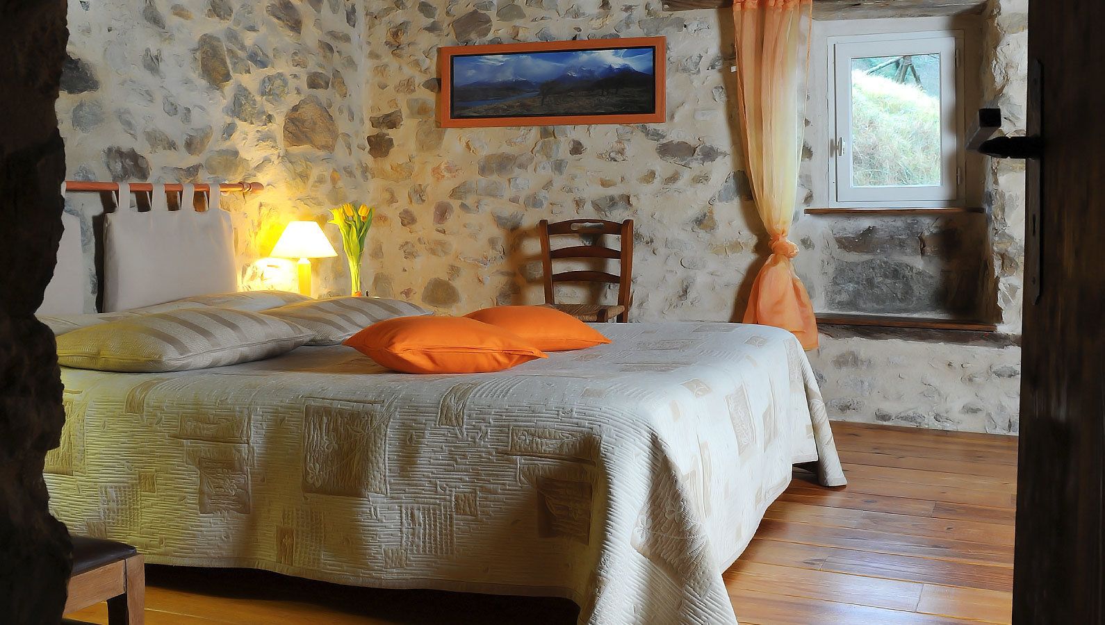 Ein Schlafzimmer mit Steinwand, einem Bett mit gemusterten Betttextilien, einem Nachttisch mit Lampe und einem Fenster mit Vorhängen.
