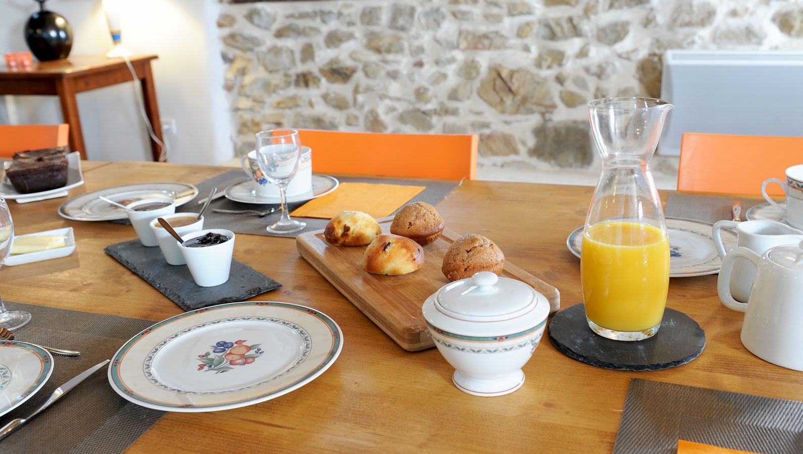 A breakfast table set with plates, a glass pitcher of orange juice, a small bowl of jam, and a wooden board with muffins.