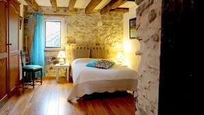 A rustic bedroom with a stone wall, wooden beams, a bed with a white bedspread and a blue pillow, a chair, and two bedside lamps. - Chambres d'Hôtes Idiartekoborda (Saint-Etienne-de-Baigorry)