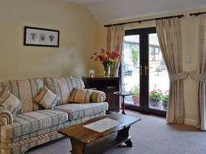 A living room with a patterned sofa, a wooden coffee table, and a door leading to an outdoor area with potted plants. - Wee Cordorcan - UK5890 (Wood of Cree, near Newton Stewart)