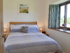 Een net opgemaakt bed met gestreepte beddengoed en hartvormige kussens, een bedlampje, een klok en een raam met gordijnen.