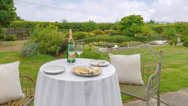 Et rundt bord med hvid dug, en champagneflaske, to vinglas, en tallerken med kiks og ost, og en bænk med metalstel og hynder.