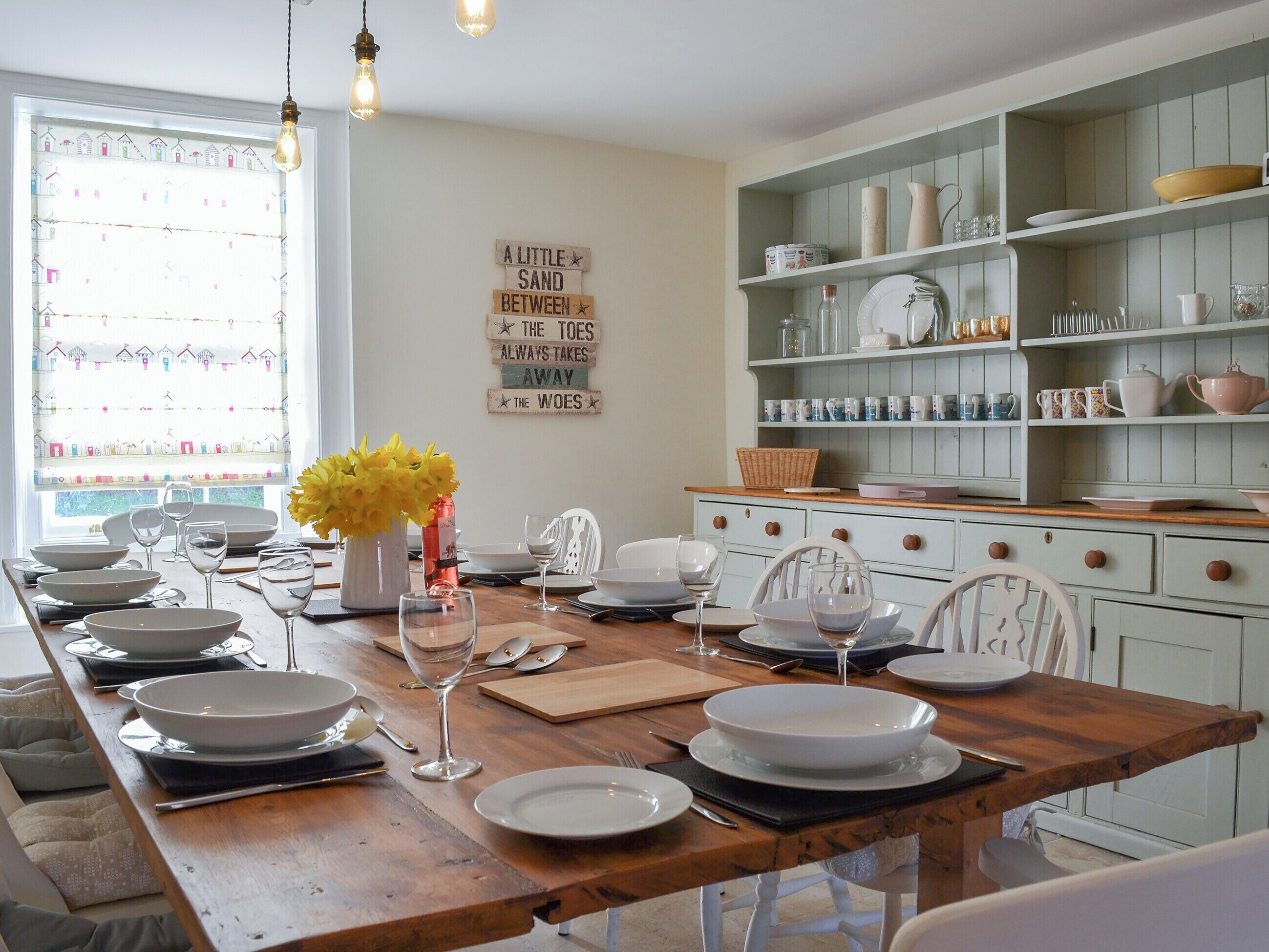A dining area with a wooden table set for six, surrounded by white chairs. A decorative sign is on the wall, and there are shelves with dishes and glassware.