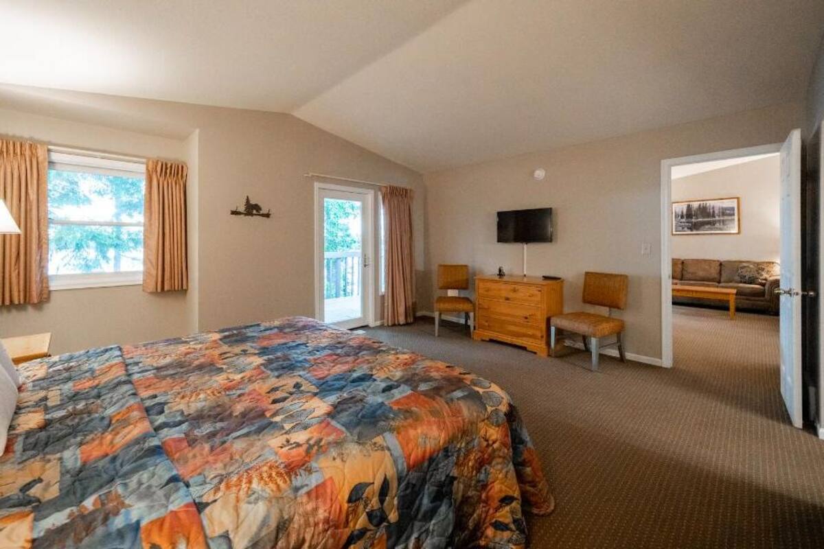 A bedroom with a large bed, a dresser, a TV, and a door leading to a living area.