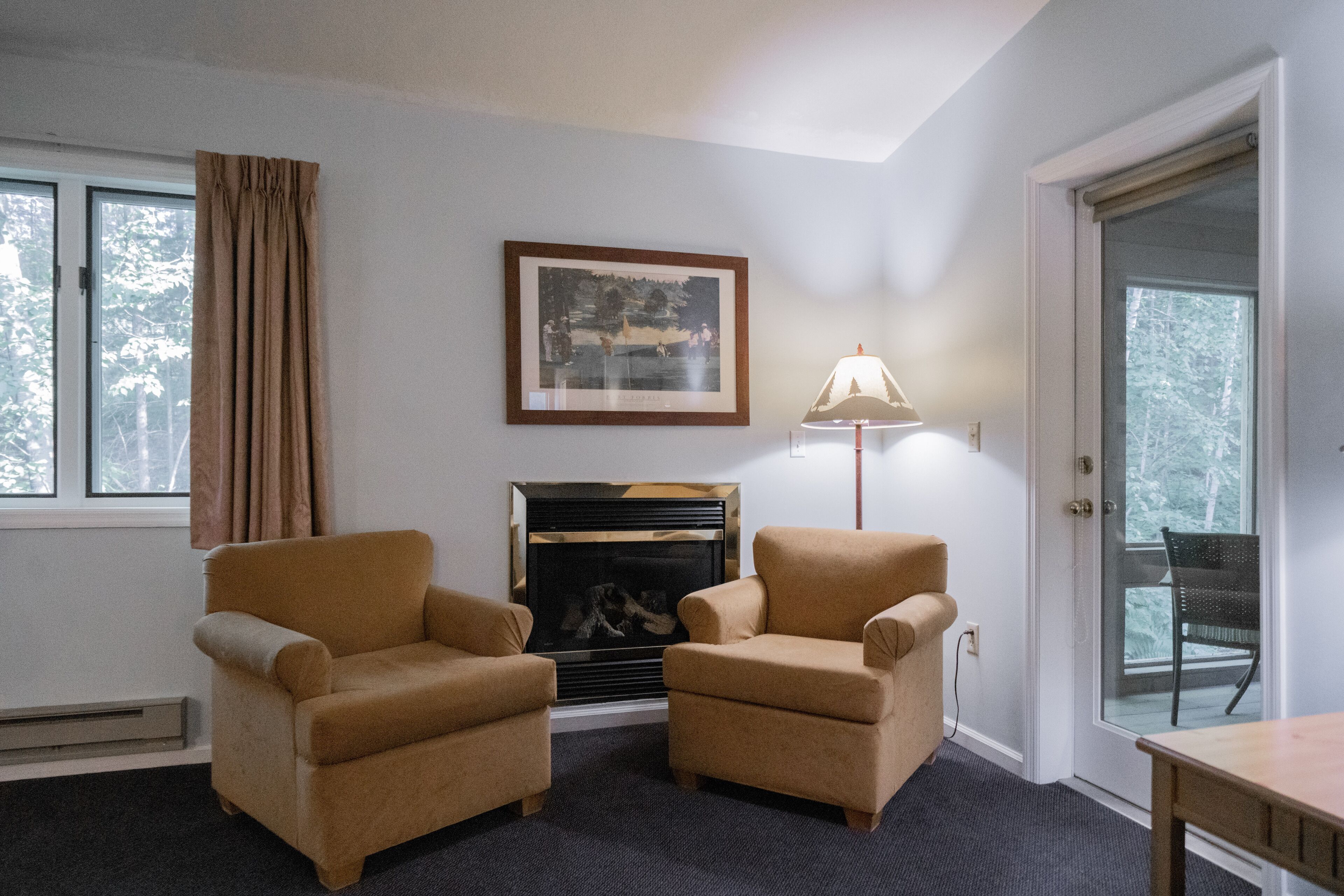 Une pièce comprenant deux fauteuils, une cheminée, un tableau encadré au mur, une lampe et une porte donnant sur un balcon.