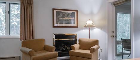 A room with two armchairs, a fireplace, a framed picture on the wall, a lamp, and a door leading to a balcony.