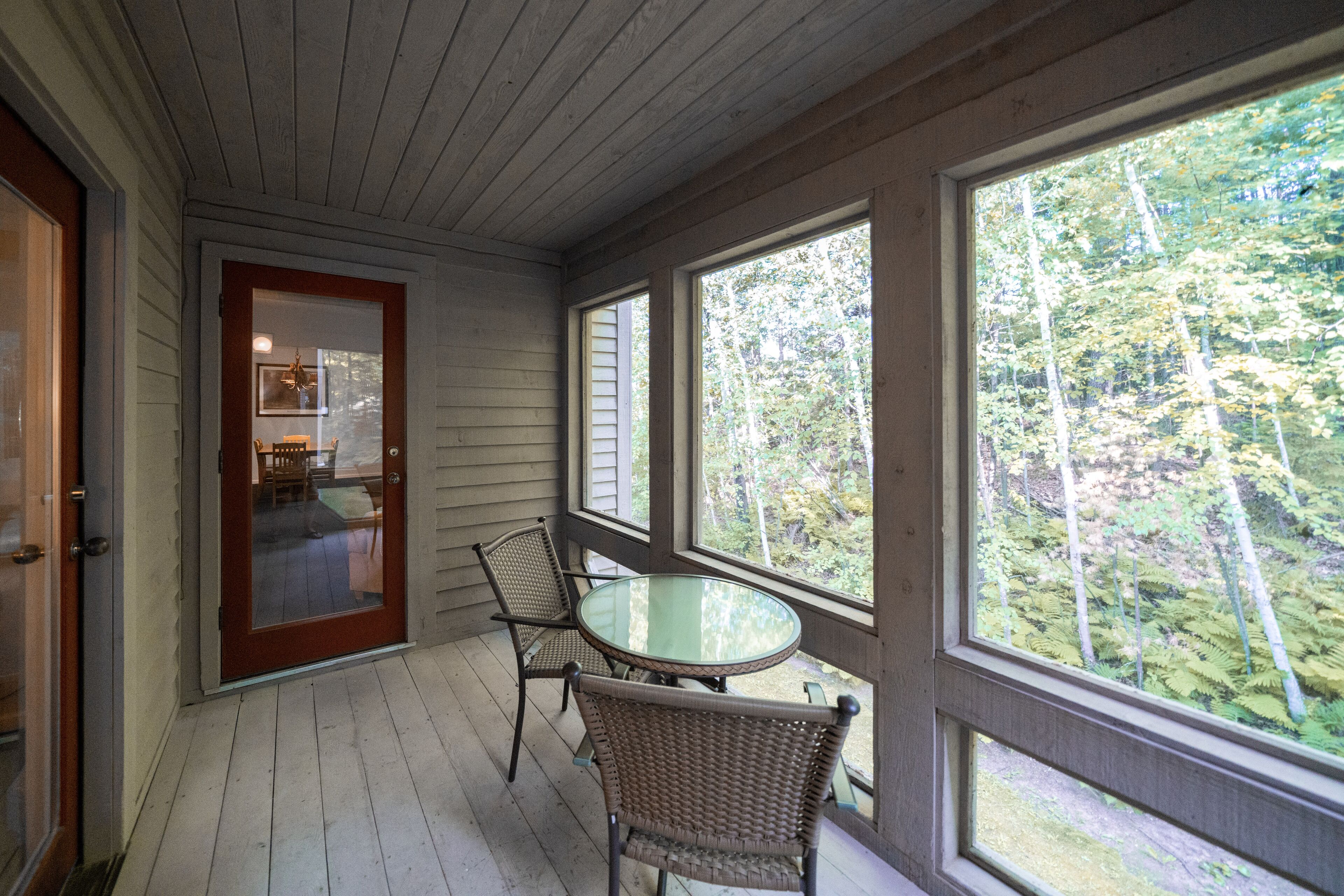 A covered porch with a glass table and wicker chairs, a wooden door, and large windows overlooking a forest.