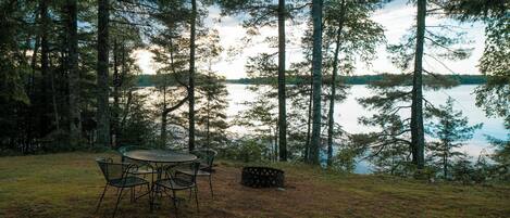 A camping area with a table and chairs, surrounded by tall trees and a lake.