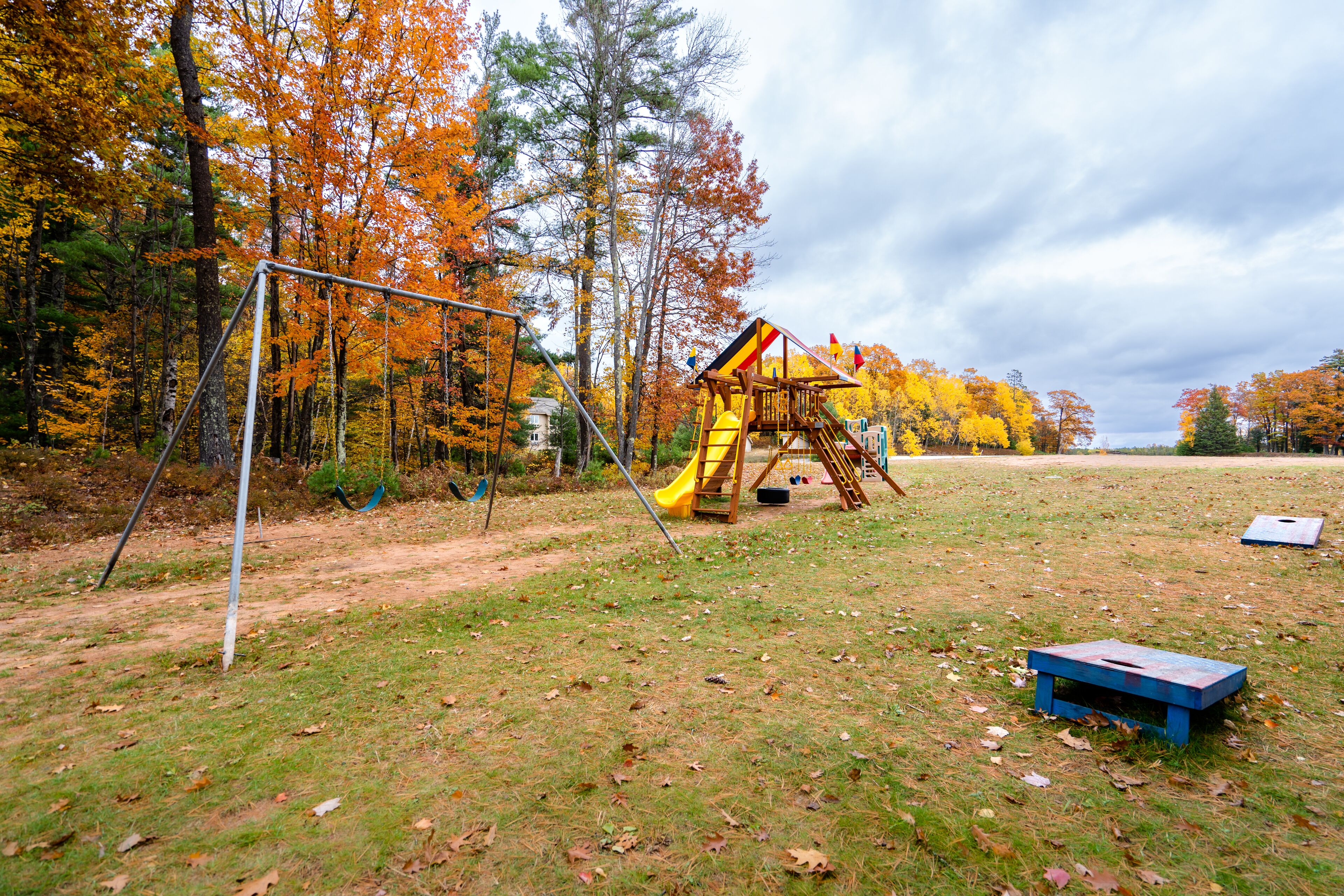 Un parque infantil con un tobogán amarillo y un banco azul en un área cubierta de césped, rodeado de árboles con follaje otoñal.
