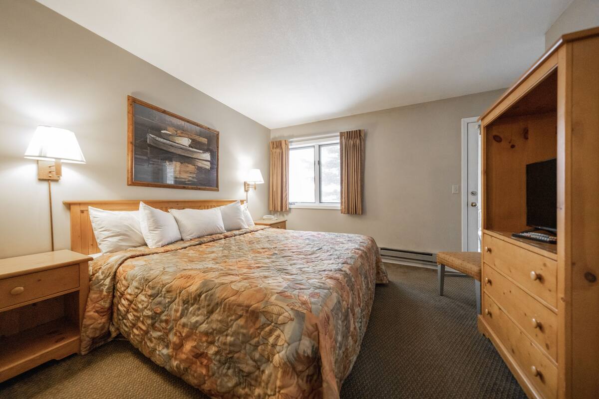 A hotel room with a large bed, wooden nightstands, a dresser, a bench, a painting on the wall, and a window with curtains.
