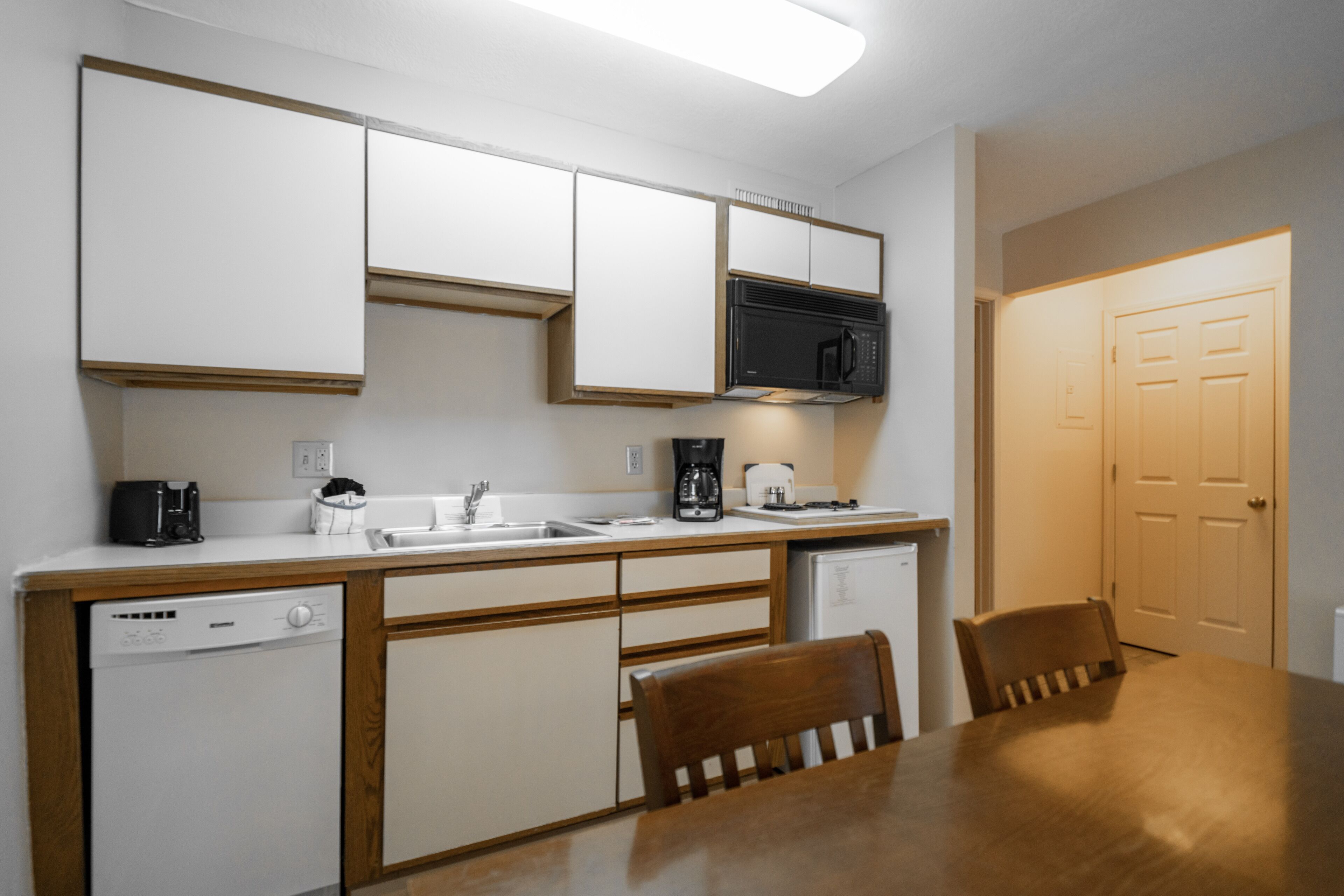 A compact kitchen with white cabinets, a microwave, a coffee maker, and a dishwasher. There is a wooden dining table with two chairs.