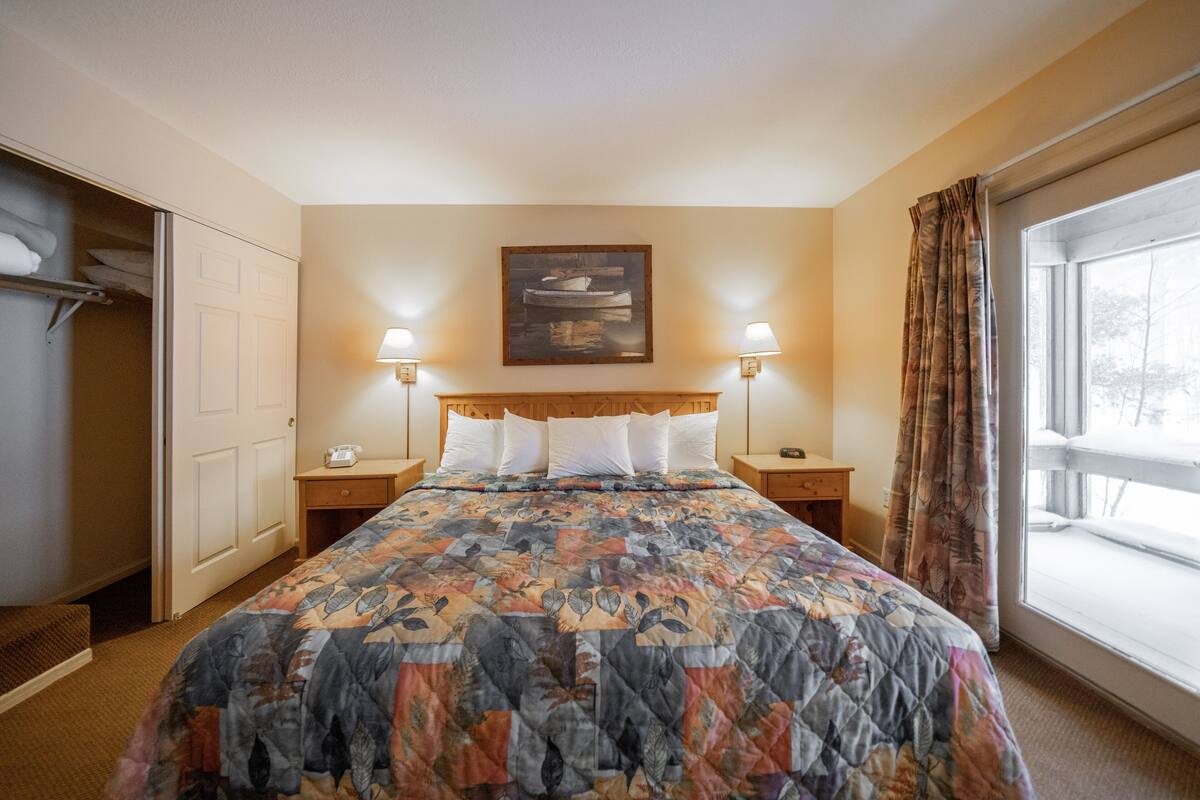 A hotel room with a large bed, a wooden headboard, two bedside lamps, a nightstand, a window with curtains, and a closet with hanging towels.
