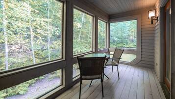 A wooden deck with large windows offering a view of a forest. There are two wicker chairs and a table.