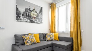 Un sofá seccional gris con cojines estampados, un cuadro de un paisaje urbano en la pared y una ventana con cortinas amarillas.