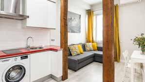 Una cocina moderna con una lavadora Bosch, un sofá gris con cojines estampados, y una zona de comedor con una mesa y sillas blancas.