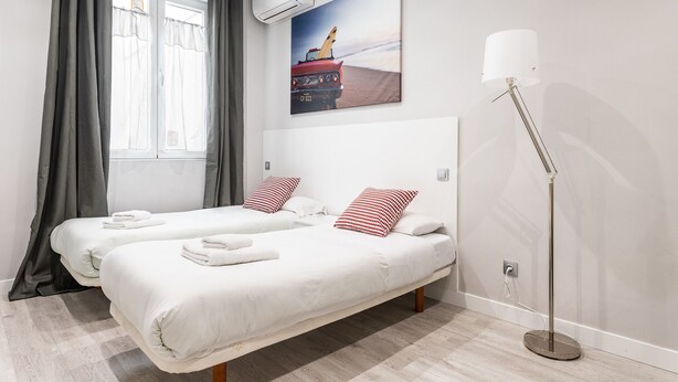 Habitación de hotel con dos camas, un ventanal con cortinas, un cuadro de un coche en la playa y una lámpara de pie.