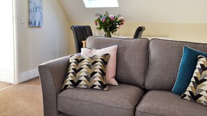 Ein graues Sofa mit gemusterten Kissen, ein schwarzer Sessel, ein kleiner Tisch mit einer Blumenvase und ein Oberlicht darüber.