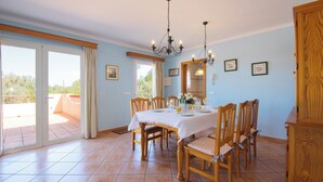 A dining area with a table set for six, wooden chairs, a chandelier, and a view through a sliding glass door to an outdoor patio. - Es figueral den Vicenç (Campos)