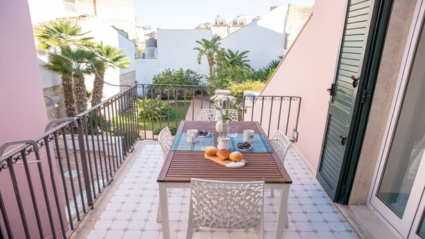 A balcony with a table set for breakfast, including croissants and a pitcher of water, surrounded by a metal railing and a view of the neighborhood. - Talè Talè Apartment just 90m from the sandy beach (Marina di Ragusa)