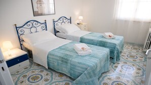 A room with two beds, each with a blue headboard and white bedding. There is a nightstand with a lamp and a small drawer. The floor has a patterned tile design. - Talè Talè Apartment just 90m from the sandy beach (Marina di Ragusa)
