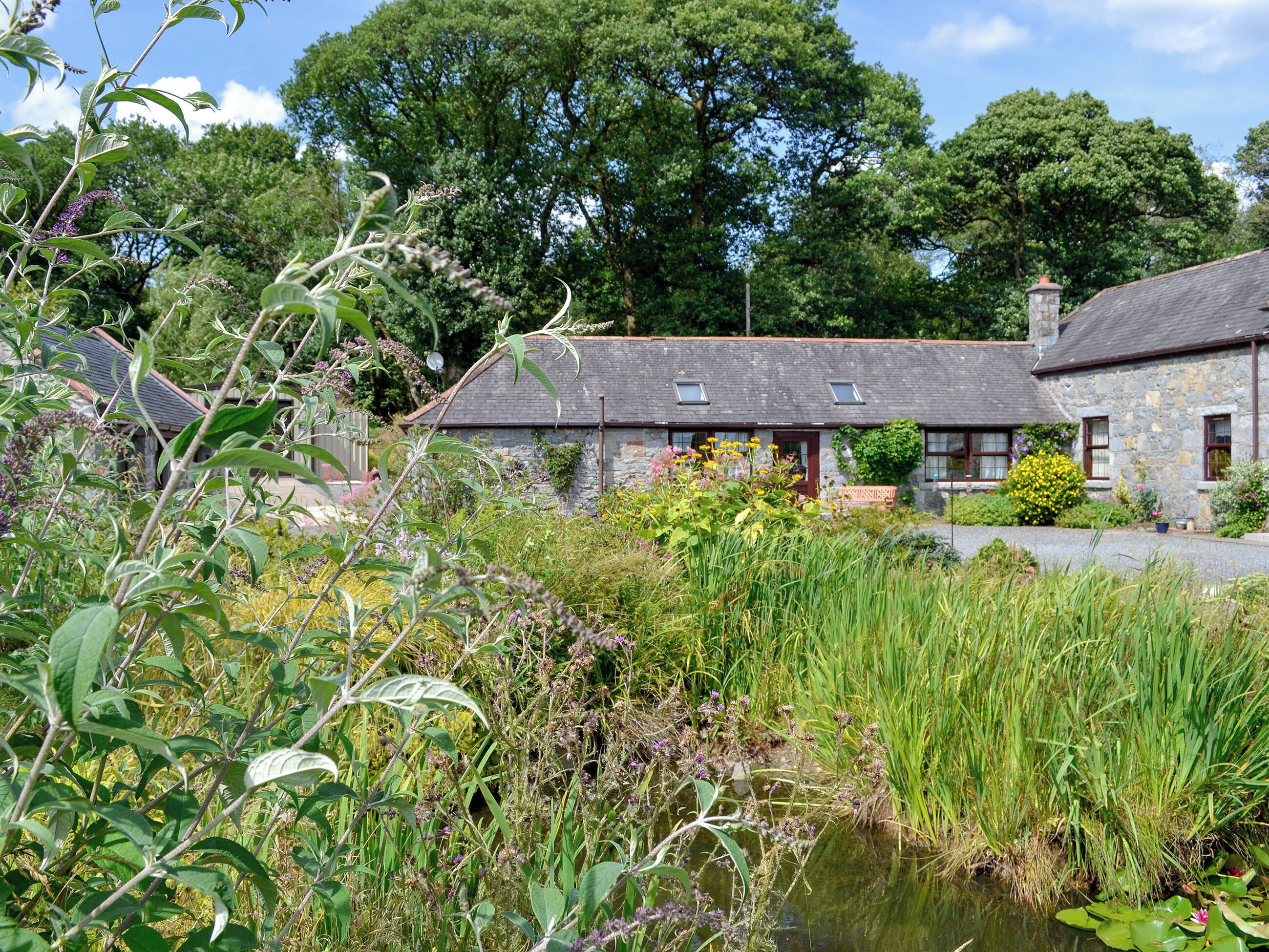 A stone cottage with a thatched roof, surrounded by lush greenery and a small pond.