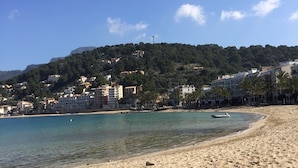 Una playa arenosa con agua azul cristalina, un bote en la orilla y edificios en una colina al fondo.
