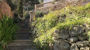 Una escalera de piedra que conduce a un edificio con balcón, rodeado de follaje y árboles.