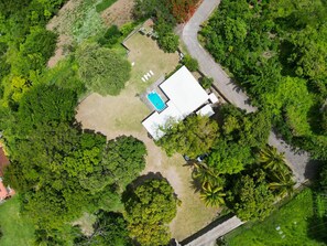 An aerial view of a property with a swimming pool, a fenced yard, and dense surrounding vegetation.