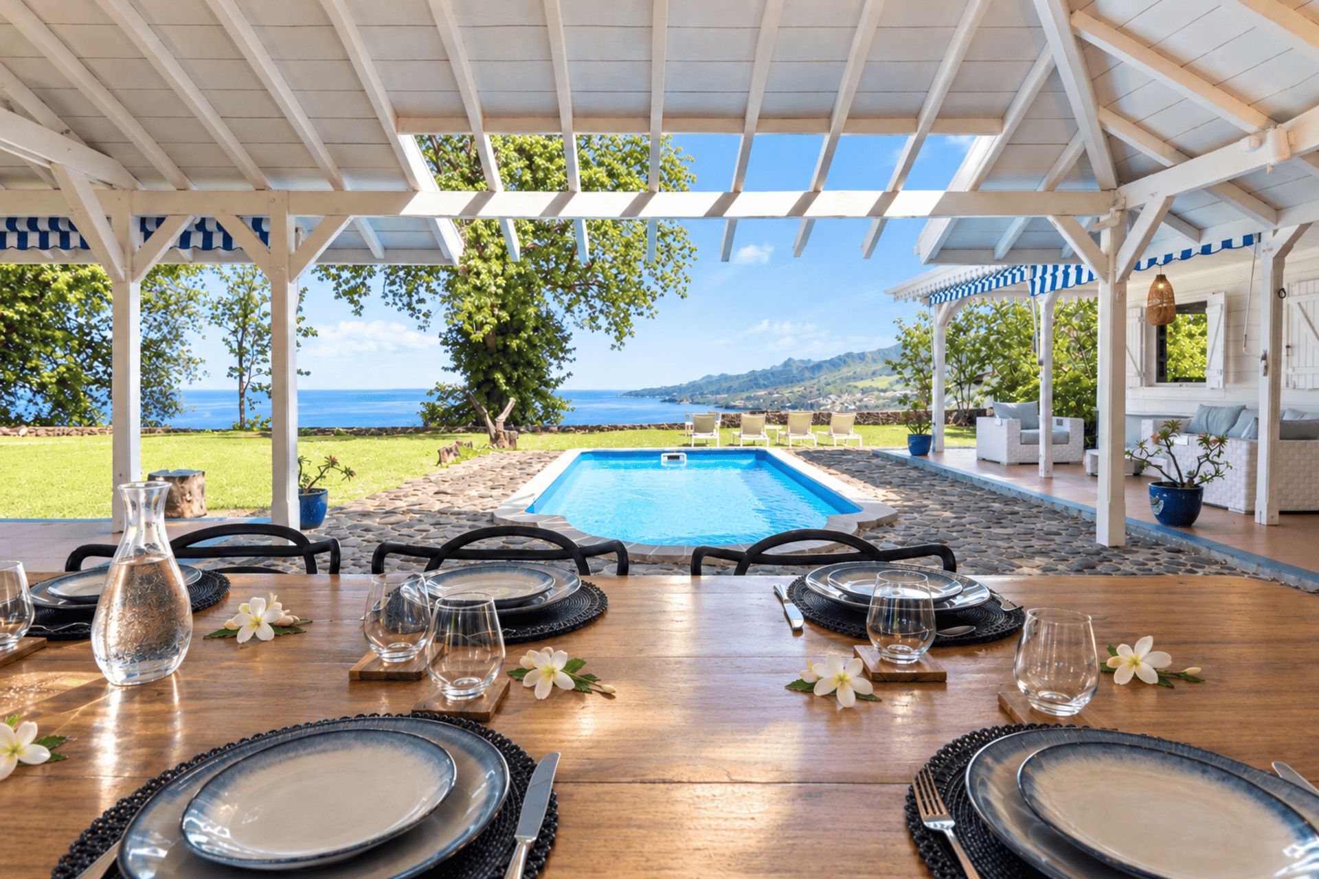 A dining area with a view of a pool and ocean, set under a pergola with a table and chairs.