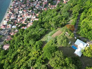An aerial view of a coastal residential area with houses, dense greenery, and a swimming pool.