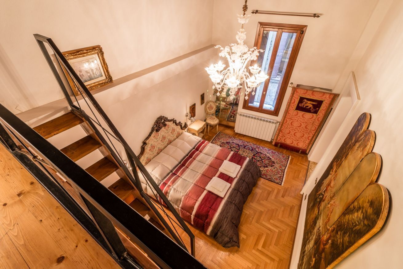 A bedroom with a wooden staircase, a bed with a red and white blanket, a chandelier, and a window with a view.