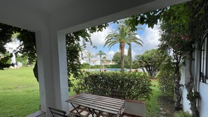 En terrasse med et bord og stole, omgivet af en have med palmer og en swimmingpool i baggrunden.