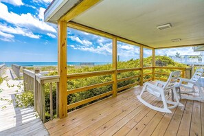 Terraza de madera con vista al océano, sillas mecedoras blancas y un área cubierta con barandilla.