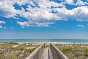 Ein Holzsteg führt zu einem Strand mit blauem Himmel und Meerblick.