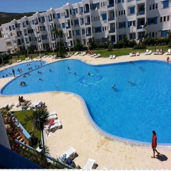 En stor swimmingpool omgiven av en sandstrand och solstolar, med en hög byggnad i bakgrunden.