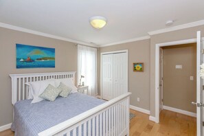 Een slaapkamer met een bed, nachtkastje, schilderij, deur en een plafondlamp.