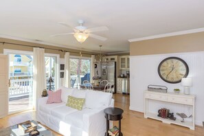 Un salon avec un canapé blanc, un ventilateur de plafond, un coin repas et une cuisine à l’arrière-plan.