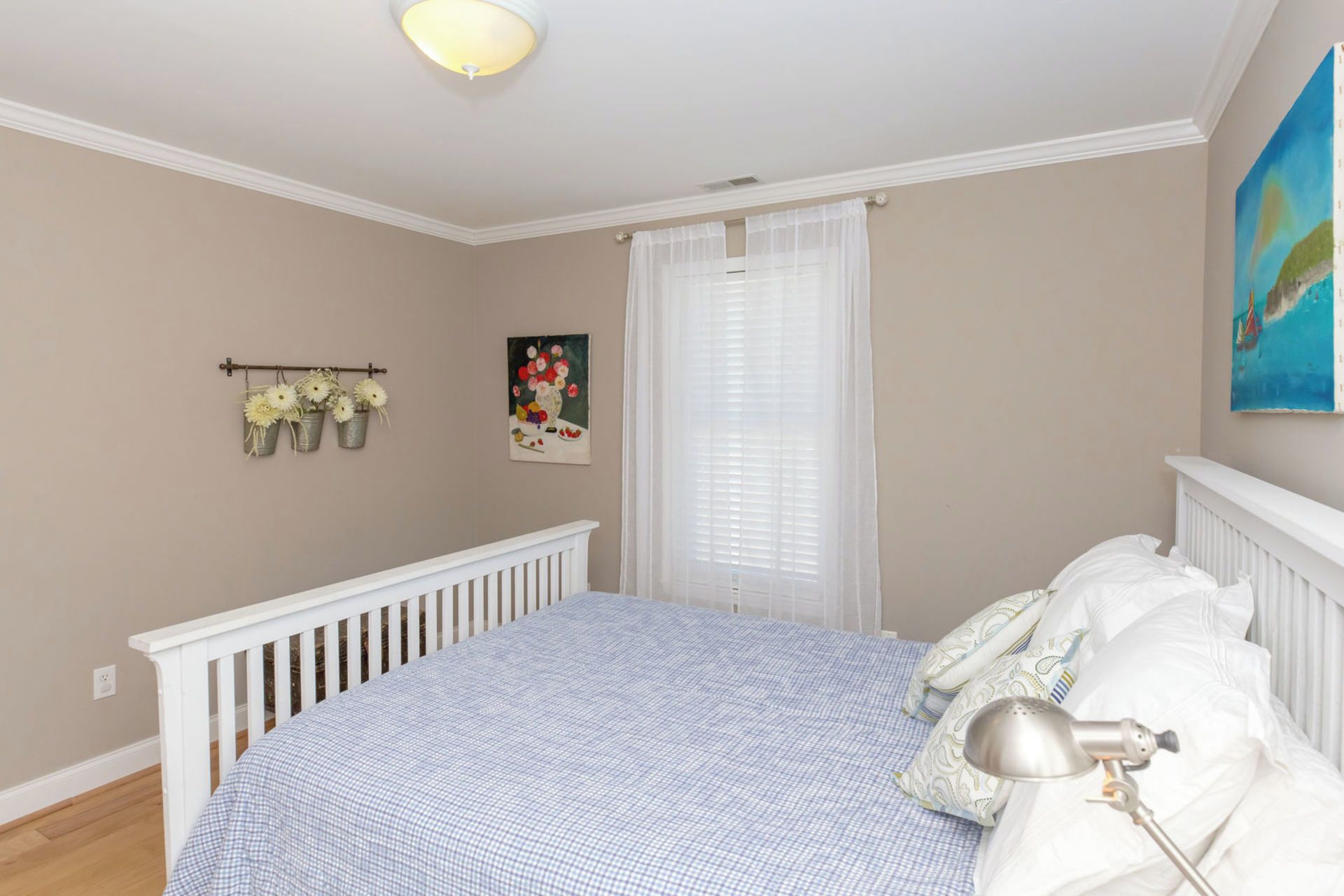 A bedroom with a white crib, a bed with a blue checkered bedspread, a white lamp, and a painting on the wall.