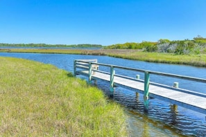 A wooden boardwalk over a calm water body, surrounded by grassy areas and trees.