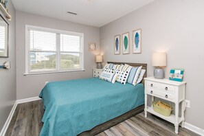A bedroom with a bed, a nightstand, a lamp, a window, and a wall with framed pictures. - Legacy Beach House - Gorgeous 4BR Oceanfront Duplex! (North Topsail Beach)