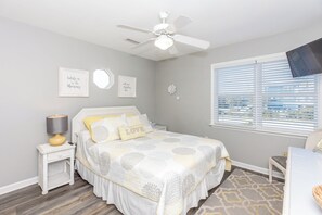 A bedroom with a bed, a nightstand, a lamp, a ceiling fan, a window with blinds, and wall decorations.