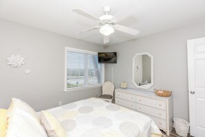 Un dormitorio con cama, cómoda, espejo, ventilador de techo y ventana con persianas.